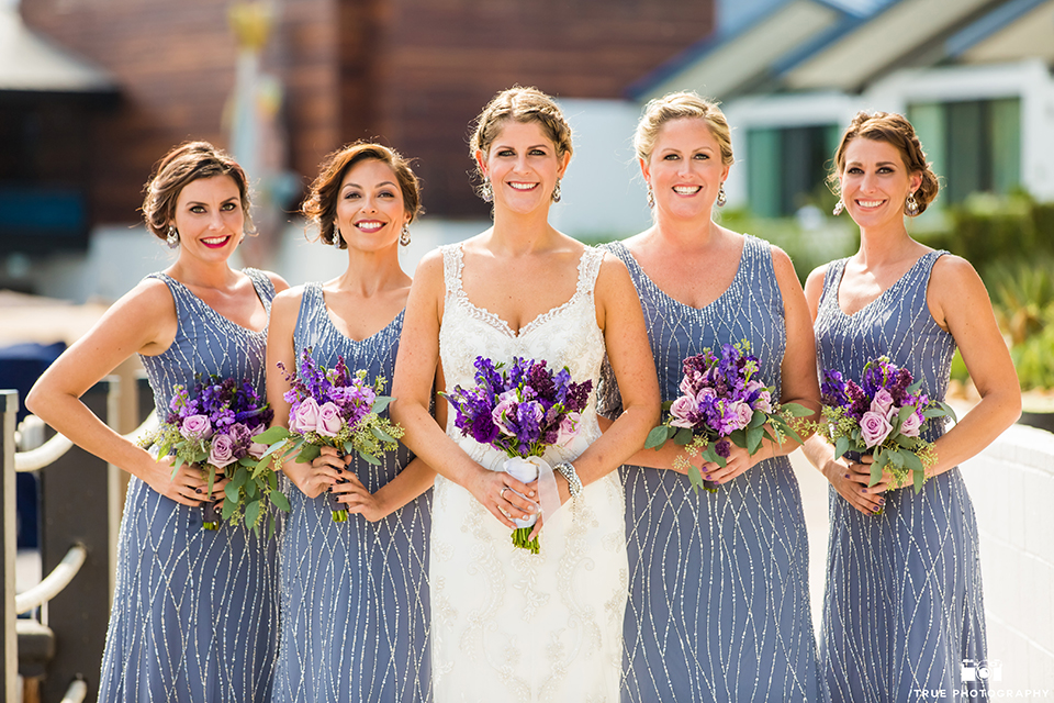 San diego outdoor wedding at leo carillo ranch bride form fitting gown with a sweetheart neckline with thin straps and beaded detail on bodice holding purple floral bridal bouquet standing with bridesmaids long lavender dresses with beaded detail holding purple floral bridal bouquets