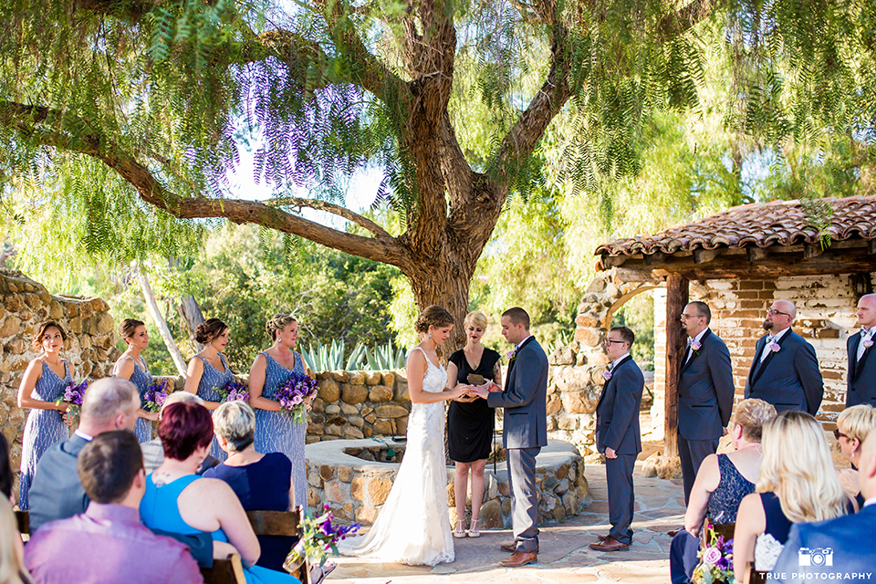 San diego outdoor wedding at leo carillo ranch bride form fitting gown with a sweetheart neckline with thin straps and beaded detail on bodice with groom charcoal grey tuxedo with a black shawl and matching vest with a white dress shirt and plaid bow tie with a white floral boutonniere holding hands during ceremony