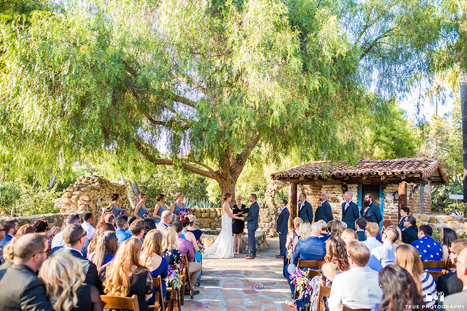 San diego outdoor wedding at leo carillo ranch bride form fitting gown with a sweetheart neckline with thin straps and beaded detail on bodice with groom charcoal grey tuxedo with a black shawl and matching vest with a white dress shirt and plaid bow tie with a white floral boutonniere holding hands during ceremony