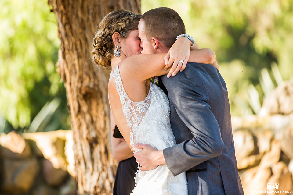 San diego outdoor wedding at leo carillo ranch bride form fitting gown with a sweetheart neckline with thin straps and beaded detail on bodice with groom charcoal grey tuxedo with a black shawl and matching vest with a white dress shirt and plaid bow tie with a white floral boutonniere kissing during ceremony