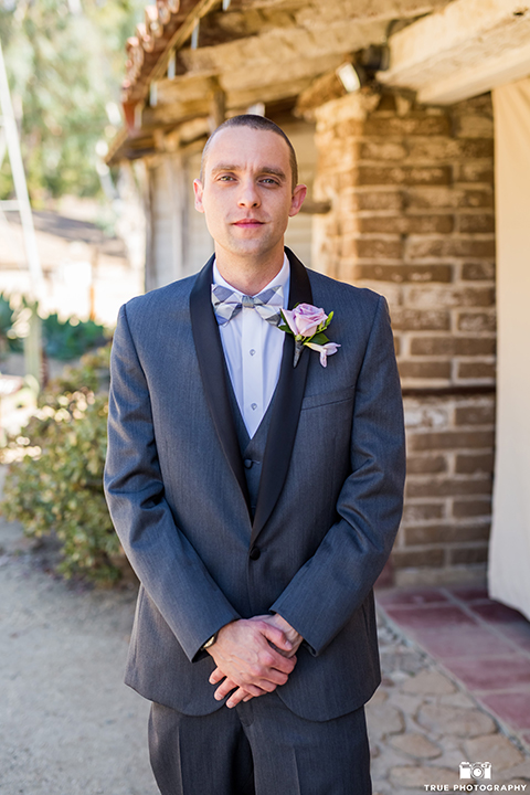 San diego outdoor wedding at leo carillo ranch groom charcoal grey tuxedo with a black shawl lapel and matching vest with a white dress shirt and purple plaid bow tie with a light purple floral boutonniere standing