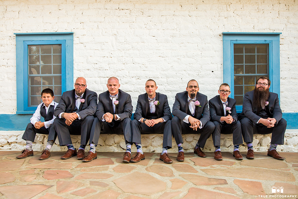 San diego outdoor wedding at leo carillo ranch groom charcoal grey tuxedo with a black shawl lapel and matching vest with a white dress shirt and purple plaid bow tie with a light purple floral boutonniere sitting with groomsmen charcoal grey tuxedos with light purple plaid bow ties