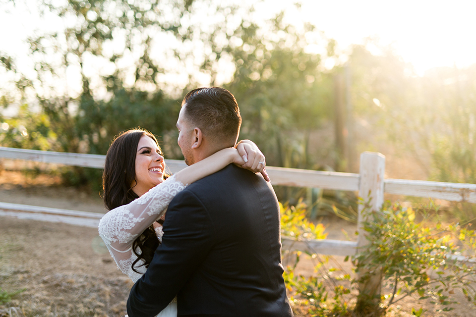 Temecula outdoor wedding at bella gardens estates bride lace ball gown with long sleeves and a sweetheart neckline with a tulle skirt and groom navy shawl lapel tuxedo with a matching vest and long navy blue tie with a white floral boutonniere hugging and smiling