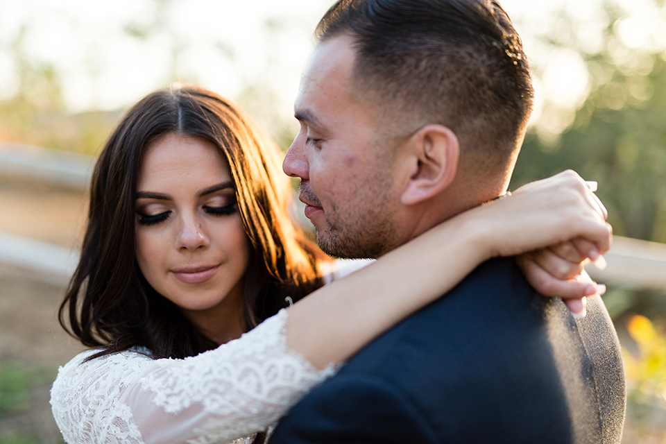 Temecula outdoor wedding at bella gardens estates bride lace ball gown with long sleeves and a sweetheart neckline with a tulle skirt and groom navy shawl lapel tuxedo with a matching vest and long navy blue tie with a white floral boutonniere hugging and smiling close up