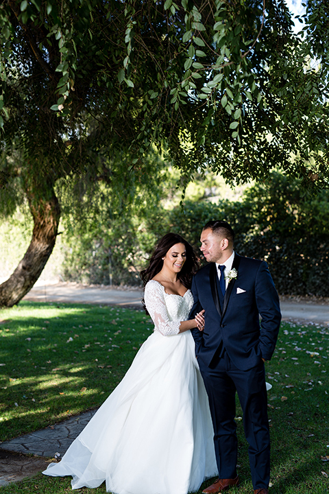 Temecula outdoor wedding at bella gardens estates bride lace ball gown with long sleeves and a sweetheart neckline with a tulle skirt and groom navy shawl lapel tuxedo with a matching vest and long navy blue tie with a white floral boutonniere standing and hugging