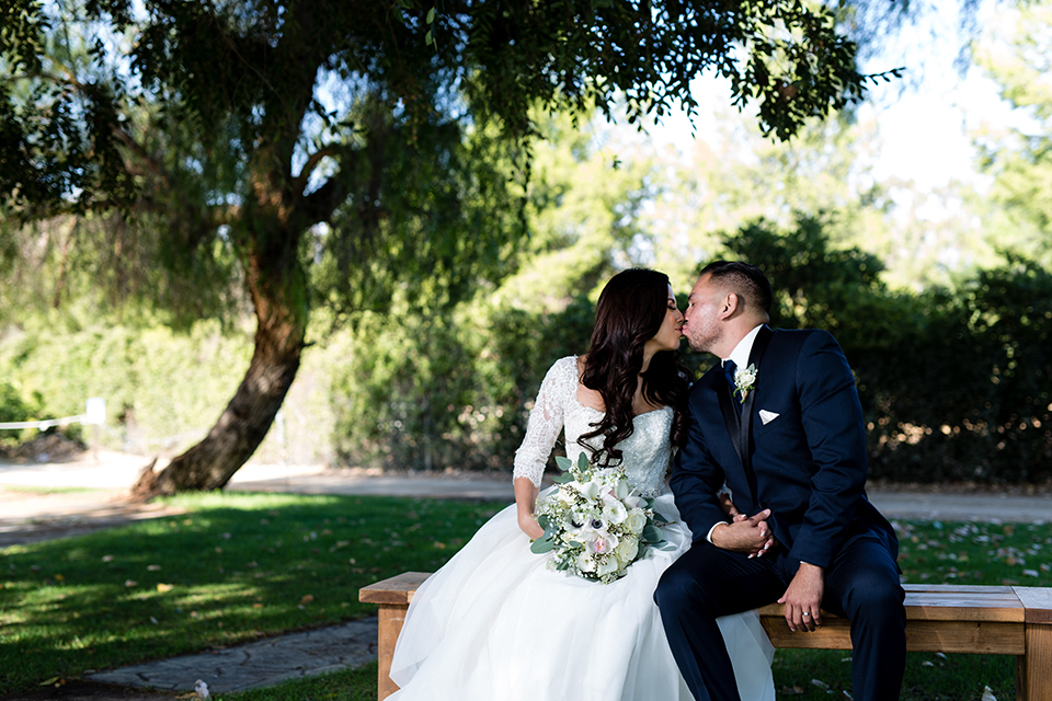 Temecula outdoor wedding at bella gardens estates bride lace ball gown with long sleeves and a sweetheart neckline with a tulle skirt and groom navy shawl lapel tuxedo with a matching vest and long navy blue tie with a white floral boutonniere sitting and kissing on bench holding white and green floral bridal bouquet