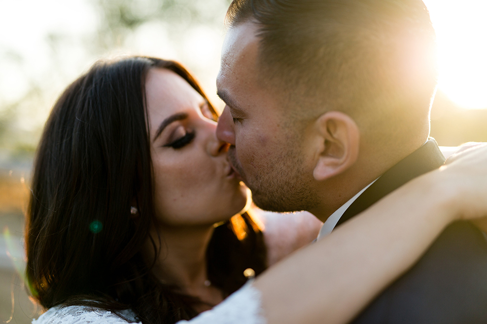 Temecula outdoor wedding at bella gardens estates bride lace ball gown with long sleeves and a sweetheart neckline with a tulle skirt and groom navy shawl lapel tuxedo with a matching vest and long navy blue tie with a white floral boutonniere kissing close up