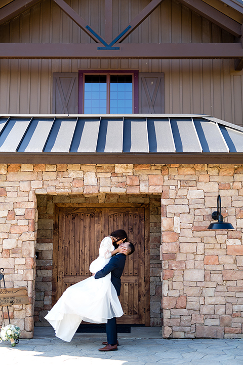 Temecula outdoor wedding at bella gardens estates bride lace ball gown with long sleeves and a sweetheart neckline with a tulle skirt and groom navy shawl lapel tuxedo with a matching vest and long navy blue tie with a white floral boutonniere hugging and kissing