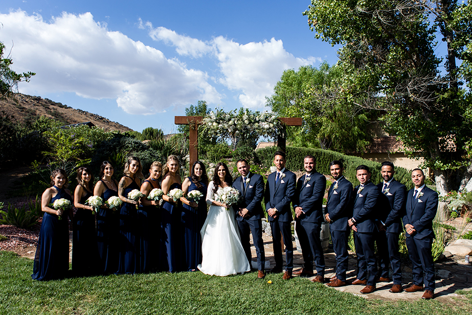 Temecula outdoor wedding at bella gardens estates bride lace ball gown with long sleeves and a sweetheart neckline with a tulle skirt and groom navy shawl lapel tuxedo with a matching vest and long navy blue tie with a white floral boutonniere with wedding party bridesmaids long blue dresses and groomsmen navy tuxedos with long ties