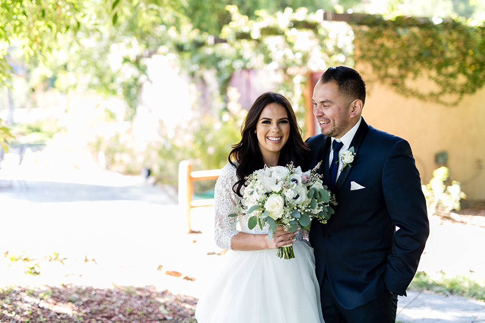 Temecula outdoor wedding at bella gardens estates bride lace ball gown with long sleeves and a sweetheart neckline with a tulle skirt and groom navy shawl lapel tuxedo with a matching vest and long navy blue tie with a white floral boutonniere hugging and bride holding white and green floral bridal bouquet