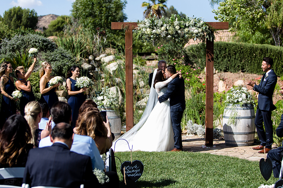 Temecula outdoor wedding at bella gardens estates bride lace ball gown with long sleeves and a sweetheart neckline with a tulle skirt and groom navy shawl lapel tuxedo with a matching vest and long navy blue tie with a white floral boutonniere kissing during ceremony