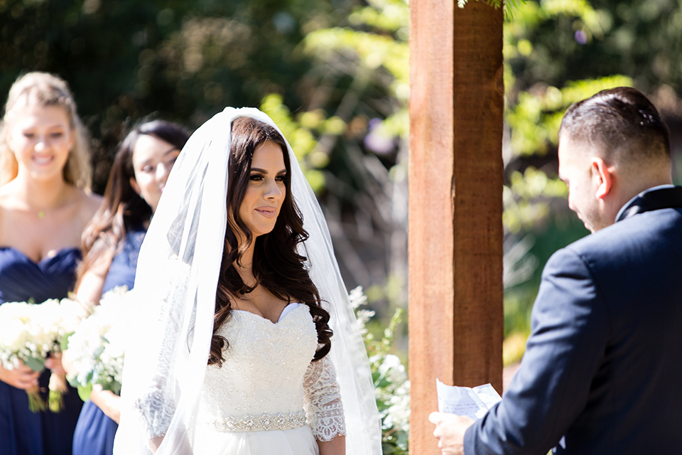 Temecula outdoor wedding at bella gardens estates bride lace ball gown with long sleeves and a sweetheart neckline with a tulle skirt and groom navy shawl lapel tuxedo with a matching vest and long navy blue tie with a white floral boutonniere smiling and holding hands during ceremony