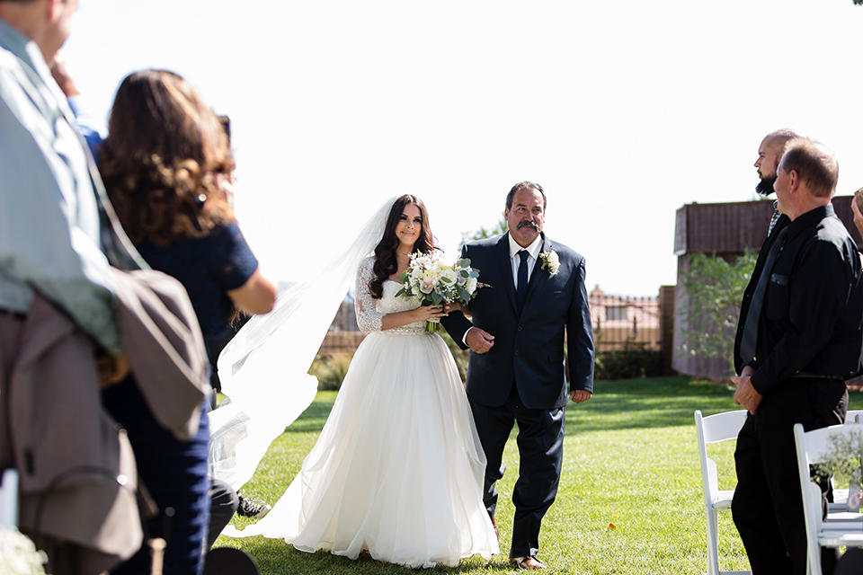 Temecula outdoor wedding at bella gardens estates bride lace ball gown with long sleeves and a sweetheart neckline with a tulle skirt holding white and green floral bridal bouquet walking down the aisle with dad