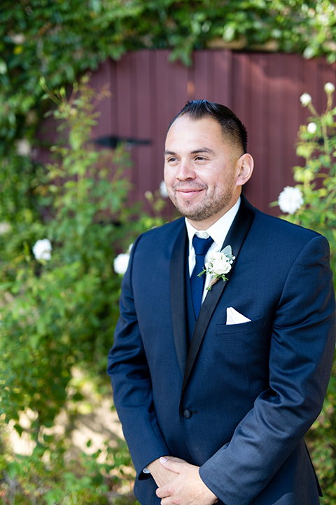 Temecula outdoor wedding at bella gardens estates groom navy shawl lapel tuxedo with a matching vest and white dress shirt with a long navy blue tie and white floral boutonniere smiling