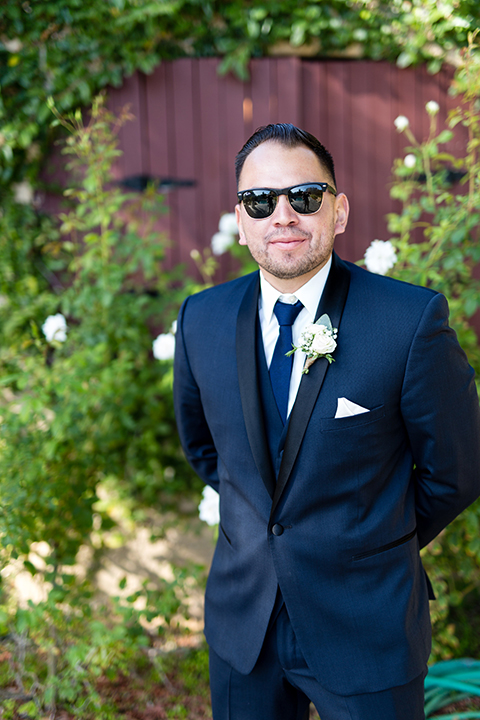 Temecula outdoor wedding at bella gardens estates groom navy shawl lapel tuxedo with a matching vest and white dress shirt with a long navy blue tie and white floral boutonniere wearing sunglasses