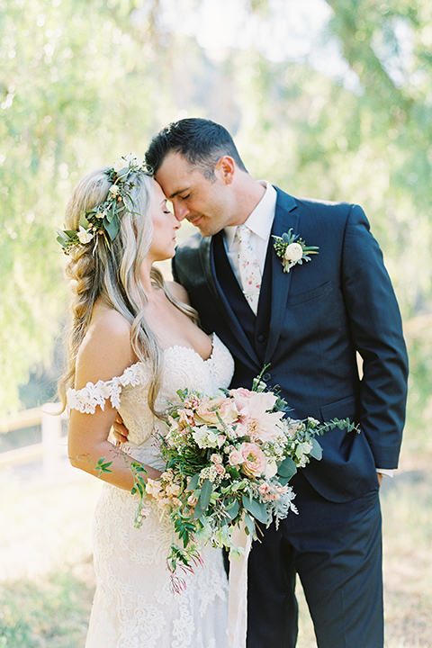 California outdoor wedding at the rancho san antonio bride form fitting lace gown with off the shoulder straps and a sweetheart neckline with a flower crown and groom navy notch lapel suit with a matching vest and white dress shirt with a long white and pink floral tie with a white floral boutonniere hugging bride holding white and green floral bridal bouquet