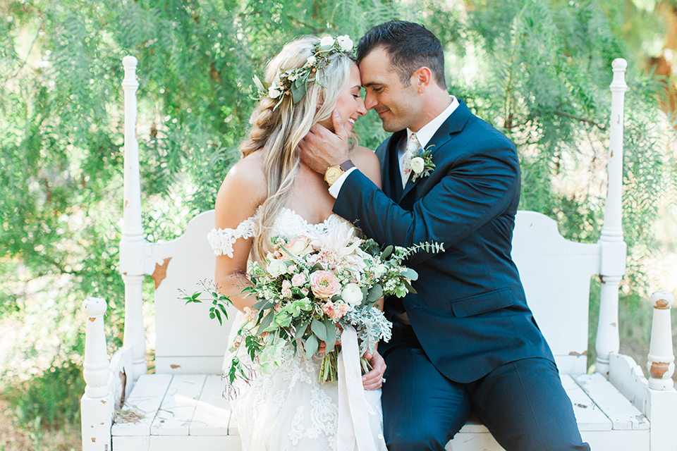 California outdoor wedding at the rancho san antonio bride form fitting lace gown with off the shoulder straps and a sweetheart neckline with a flower crown and groom navy notch lapel suit with a matching vest and white dress shirt with a long white and pink floral tie with a white floral boutonniere sitting and hugging bride holding white and green floral bridal bouquet