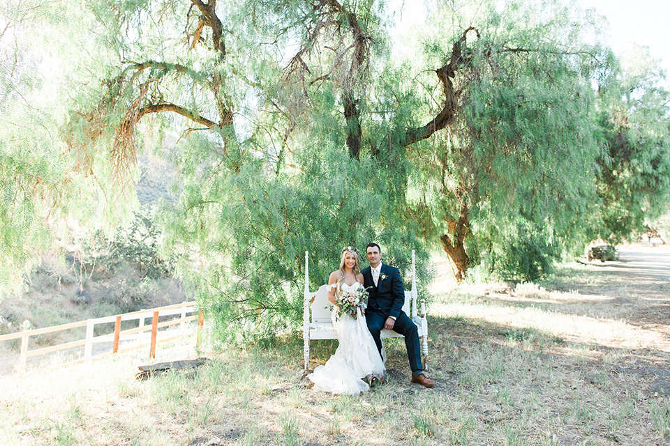 California outdoor wedding at the rancho san antonio bride form fitting lace gown with off the shoulder straps and a sweetheart neckline with a flower crown and groom navy notch lapel suit with a matching vest and white dress shirt with a long white and pink floral tie with a white floral boutonniere sitting and bride holding white and green floral bridal bouquet