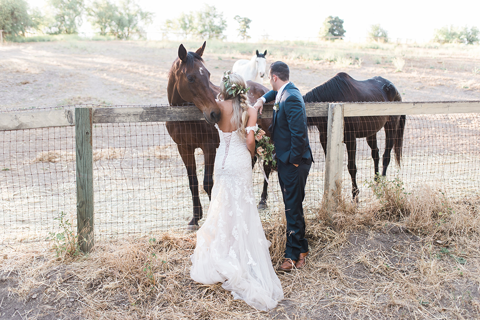 California outdoor wedding at the rancho san antonio bride form fitting lace gown with off the shoulder straps and a sweetheart neckline with a flower crown and groom navy notch lapel suit with a matching vest and white dress shirt with a long white and pink floral tie with a white floral boutonniere standing by horse bride holding white and green floral bridal bouquet