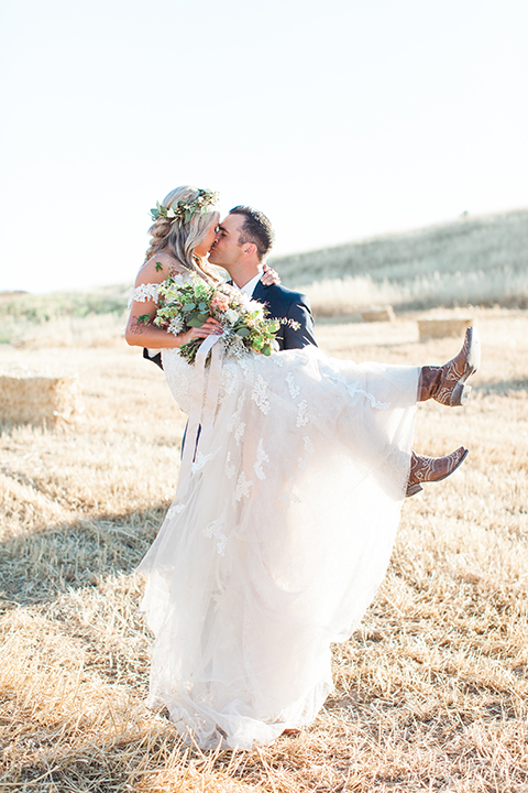 California outdoor wedding at the rancho san antonio bride form fitting lace gown with off the shoulder straps and a sweetheart neckline with a flower crown and groom navy notch lapel suit with a matching vest and white dress shirt with a long white and pink floral tie with a white floral boutonniere hugging and kissing groom carrying bride holding white and green floral bridal bouquet