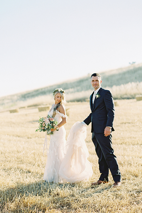 California outdoor wedding at the rancho san antonio bride form fitting lace gown with off the shoulder straps and a sweetheart neckline with a flower crown and groom navy notch lapel suit with a matching vest and white dress shirt with a long white and pink floral tie with a white floral boutonniere standing and groom holding brides dress holding white and green floral bridal bouquet