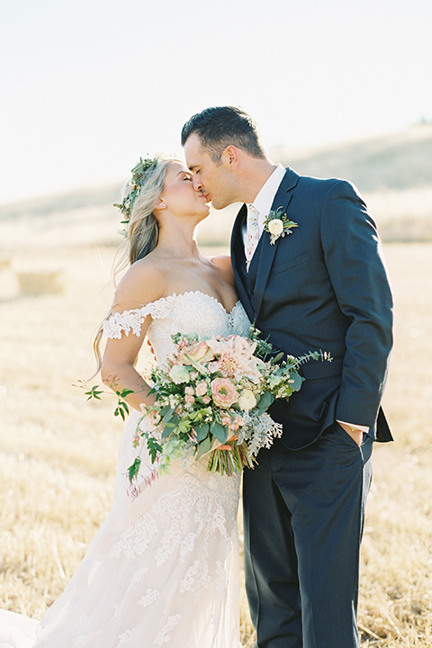 California outdoor wedding at the rancho san antonio bride form fitting lace gown with off the shoulder straps and a sweetheart neckline with a flower crown and groom navy notch lapel suit with a matching vest and white dress shirt with a long white and pink floral tie with a white floral boutonniere hugging and kissing bride holding white and green floral bridal bouquet