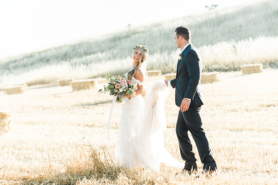 California outdoor wedding at the rancho san antonio bride form fitting lace gown with off the shoulder straps and a sweetheart neckline with a flower crown and groom navy notch lapel suit with a matching vest and white dress shirt with a long white and pink floral tie with a white floral boutonniere standing and walking groom holding dress bride holding white and green floral bridal bouquet