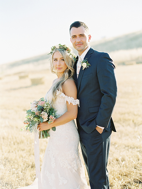 California outdoor wedding at the rancho san antonio bride form fitting lace gown with off the shoulder straps and a sweetheart neckline with a flower crown and groom navy notch lapel suit with a matching vest and white dress shirt with a long white and pink floral tie with a white floral boutonniere standing and smiling bride holding white and green floral bridal bouquet