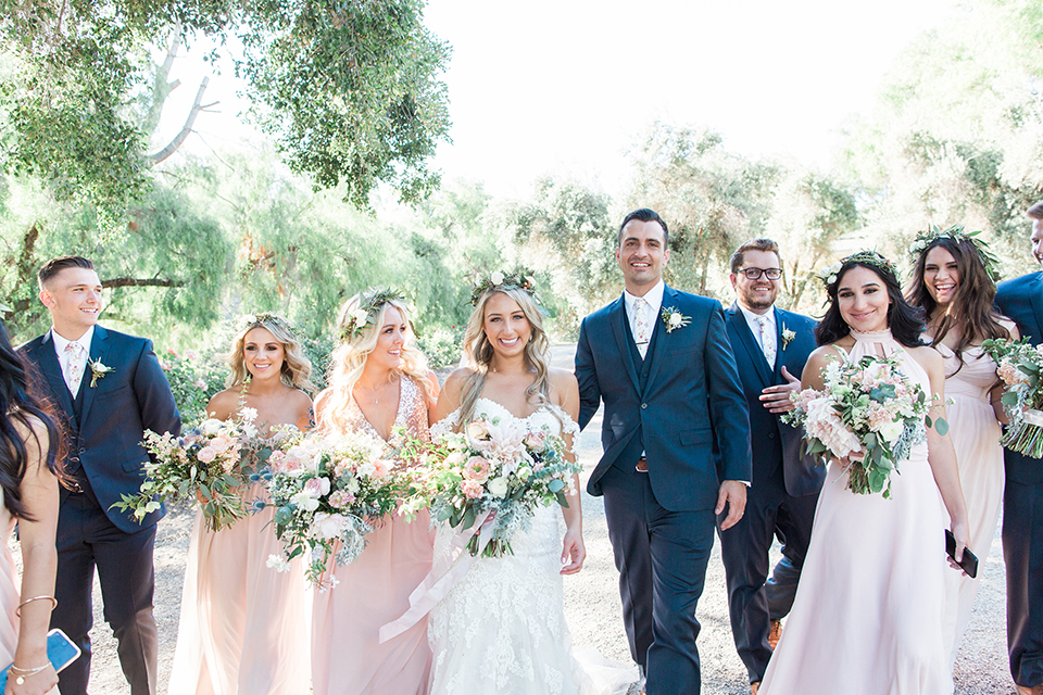 California outdoor wedding at the rancho san antonio bride form fitting lace gown with off the shoulder straps and a sweetheart neckline with a flower crown and groom navy notch lapel suit with a matching vest and white dress shirt with a long white and pink floral tie with a white floral boutonniere walking with wedding party bridesmaids long blush pink dresses and groomsmen navy suits with long floral ties