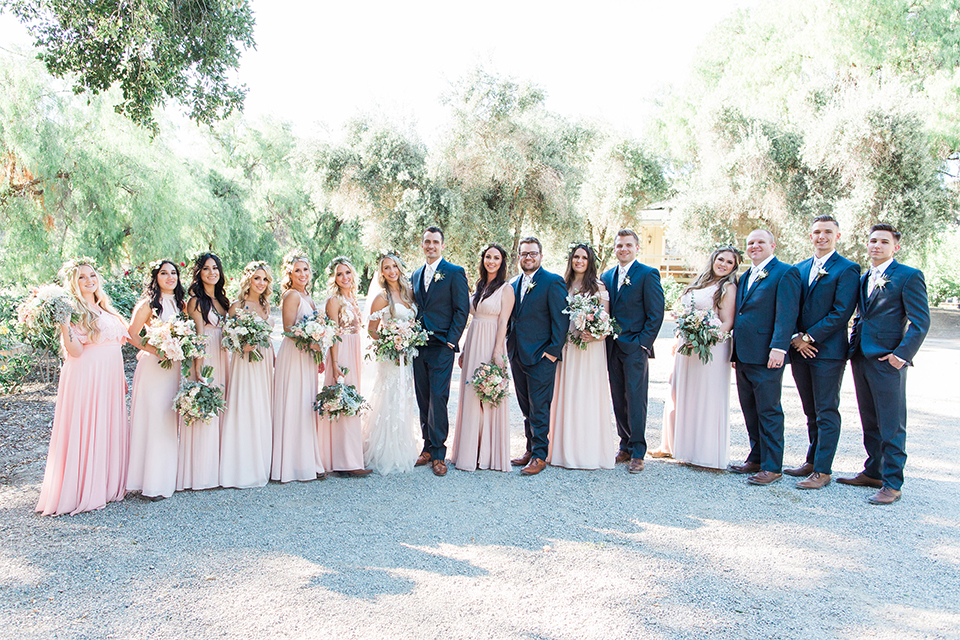 California outdoor wedding at the rancho san antonio bride form fitting lace gown with off the shoulder straps and a sweetheart neckline with a flower crown and groom navy notch lapel suit with a matching vest and white dress shirt with a long white and pink floral tie with a white floral boutonniere standing with wedding party bridesmaids long blush pink dresses and groomsmen navy blue suits with long floral ties