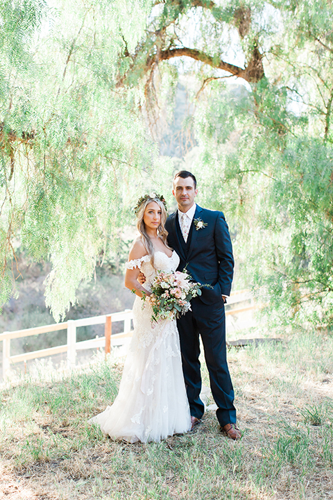 California outdoor wedding at the rancho san antonio bride form fitting lace gown with off the shoulder straps and a sweetheart neckline with a flower crown and groom navy notch lapel suit with a matching vest and white dress shirt with a long white and pink floral tie with a white floral boutonniere standing and hugging bride holding white and green floral bridal bouquet