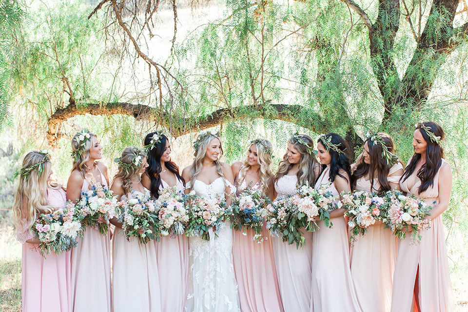 California outdoor wedding at the rancho san antonio bride form fitting lace gown with off the shoulder straps and a sweetheart neckline with a flower crown holding white and green floral bridal bouquet standing with bridesmaids long blush pink dresses and white floral bouquets