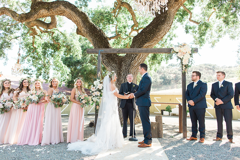 California outdoor wedding at the rancho san antonio bride form fitting lace gown with off the shoulder straps and a sweetheart neckline with a flower crown and groom navy notch lapel suit with a matching vest and white dress shirt with a long white and pink floral tie with a white floral boutonniere holding hands during ceremony