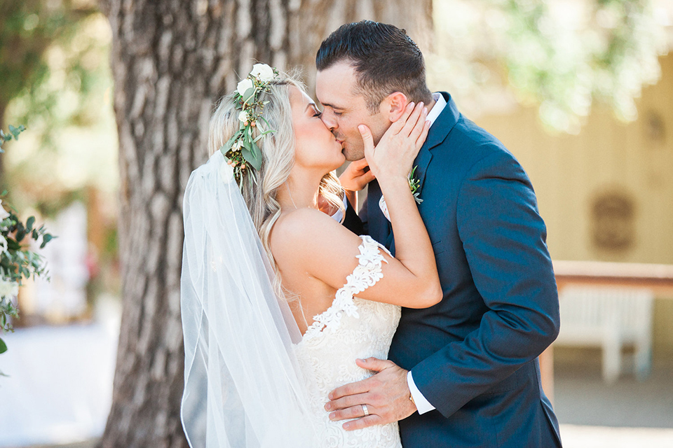 California outdoor wedding at the rancho san antonio bride form fitting lace gown with off the shoulder straps and a sweetheart neckline with a flower crown and groom navy notch lapel suit with a matching vest and white dress shirt with a long white and pink floral tie with a white floral boutonniere kissing during ceremony