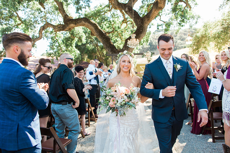 California outdoor wedding at the rancho san antonio bride form fitting lace gown with off the shoulder straps and a sweetheart neckline with a flower crown and groom navy notch lapel suit with a matching vest and white dress shirt with a long white and pink floral tie with a white floral boutonniere holding hands and walking down the aisle after ceremony bride holding white and green floral bridal bouquet