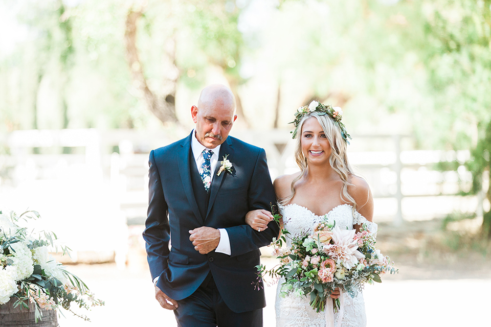 California outdoor wedding at the rancho san antonio bride form fitting lace gown with off the shoulder straps and a sweetheart neckline with a flower crown holding white and green floral bridal bouquet walking down the aisle with dad holding white and green floral bridal bouquet