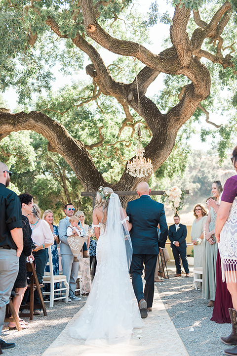 California outdoor wedding at the rancho san antonio bride form fitting lace gown with off the shoulder straps and a sweetheart neckline with a flower crown holding white and green floral bridal bouquet walking down the aisle with dad holding white and green floral bridal bouquet