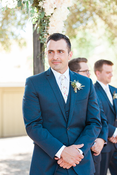 California outdoor wedding at the rancho san antonio groom navy notch lapel suit with a matching vest and white dress shirt with a long white and pink floral tie with a white floral boutonniere watching bride walk down the aisle during ceremony 