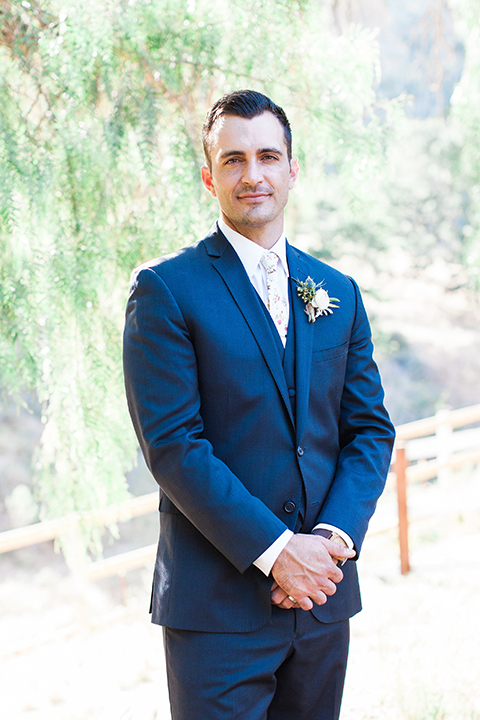 California outdoor wedding at the rancho san antonio groom navy notch lapel suit with a matching vest and white dress shirt with a long white and pink floral tie with a white floral boutonniere standing with hands folded