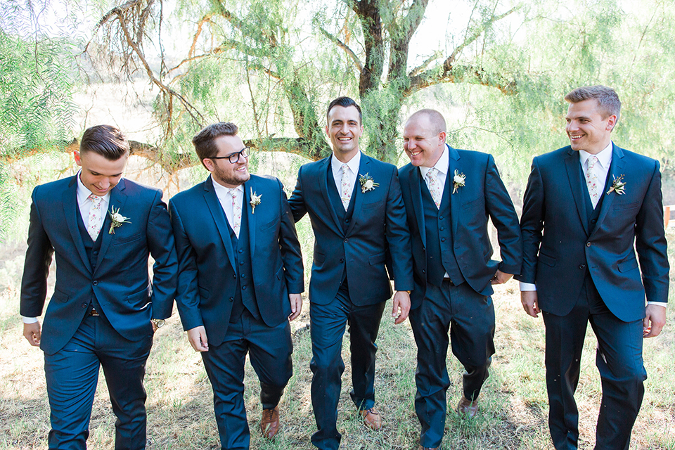 California outdoor wedding at the rancho san antonio groom navy notch lapel suit with a matching vest and white dress shirt with a long white and pink floral tie with a white floral boutonniere walking with groomsmen navy suits with long white floral ties