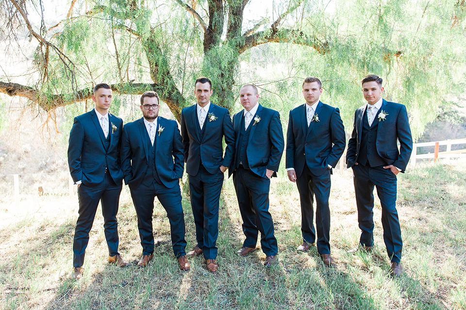 California outdoor wedding at the rancho san antonio groom navy notch lapel suit with a matching vest and white dress shirt with a long white and pink floral tie with a white floral boutonniere standing with groomsmen navy suits with long white floral ties
