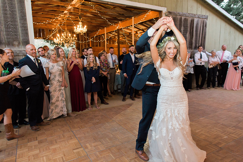 California outdoor wedding at the rancho san antonio bride form fitting lace gown with off the shoulder straps and a sweetheart neckline with a flower crown and groom navy notch lapel suit with a matching vest and white dress shirt with a long white and pink floral tie with a white floral boutonniere dancing at reception