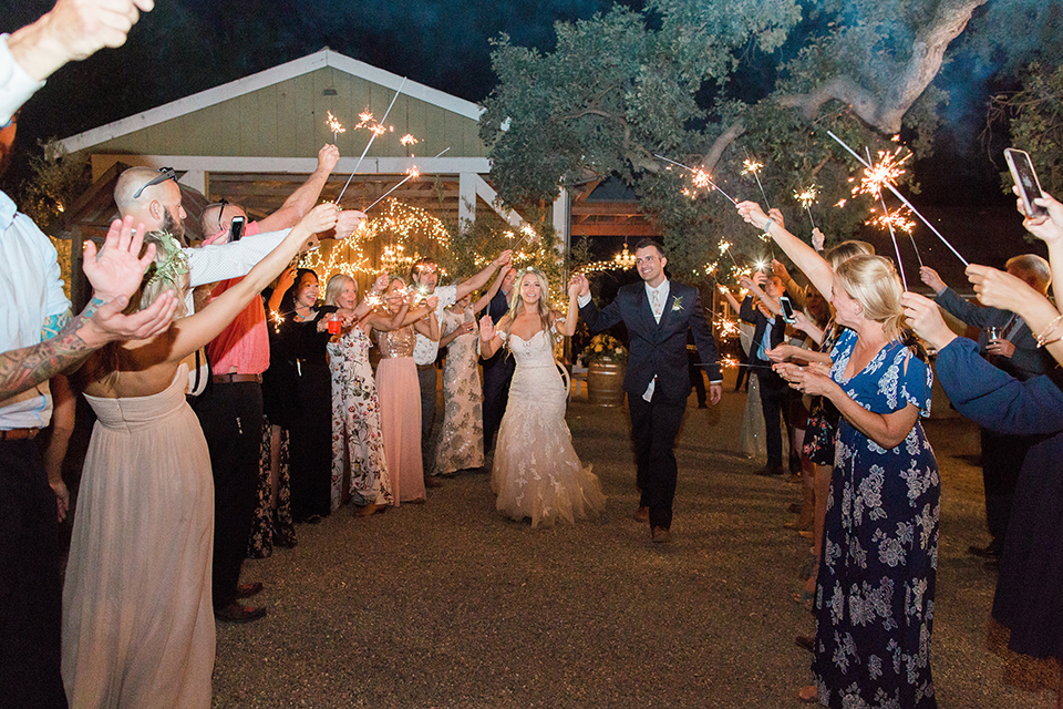 California outdoor wedding at the rancho san antonio bride form fitting lace gown with off the shoulder straps and a sweetheart neckline with a flower crown and groom navy notch lapel suit with a matching vest and white dress shirt with a long white and pink floral tie with a white floral boutonniere holding hands and walking during sparkler exit