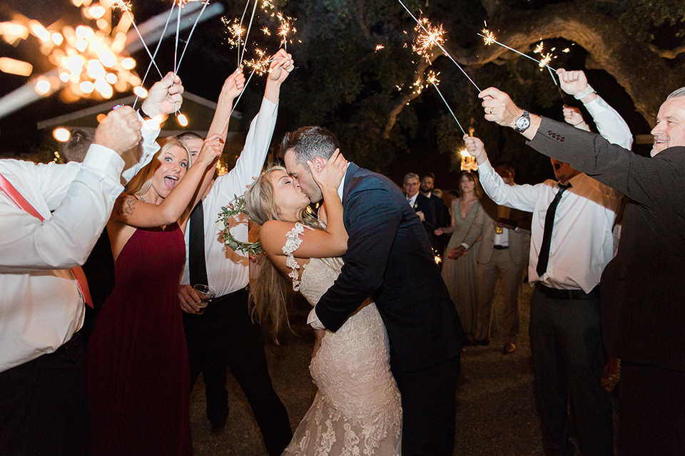 California outdoor wedding at the rancho san antonio bride form fitting lace gown with off the shoulder straps and a sweetheart neckline with a flower crown and groom navy notch lapel suit with a matching vest and white dress shirt with a long white and pink floral tie with a white floral boutonniere hugging and kissing during sparkler exit