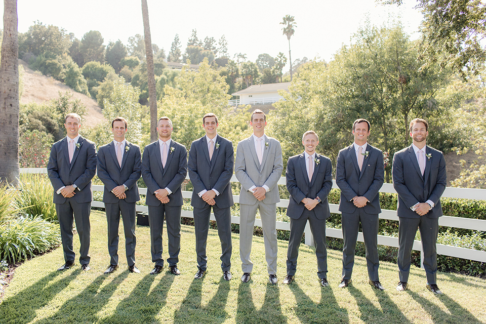 Rancho palos verdes outdoor wedding at a private estate groom heather grey notch lapel suit with a matching vest and white dress shirt with a long white tie and white floral boutonniere standing with groomsmen charcoal grey suits with long blush ties