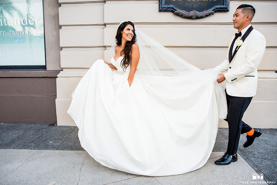 San diego glamorous wedding at the us grant hotel bride strapless ball gown with a tulle skirt and sweetheart neckline and a crystal belt with a long veil and groom white tuxedo jacket with a black shawl and black pants with a black bow tie and white floral boutonniere holding brides dress