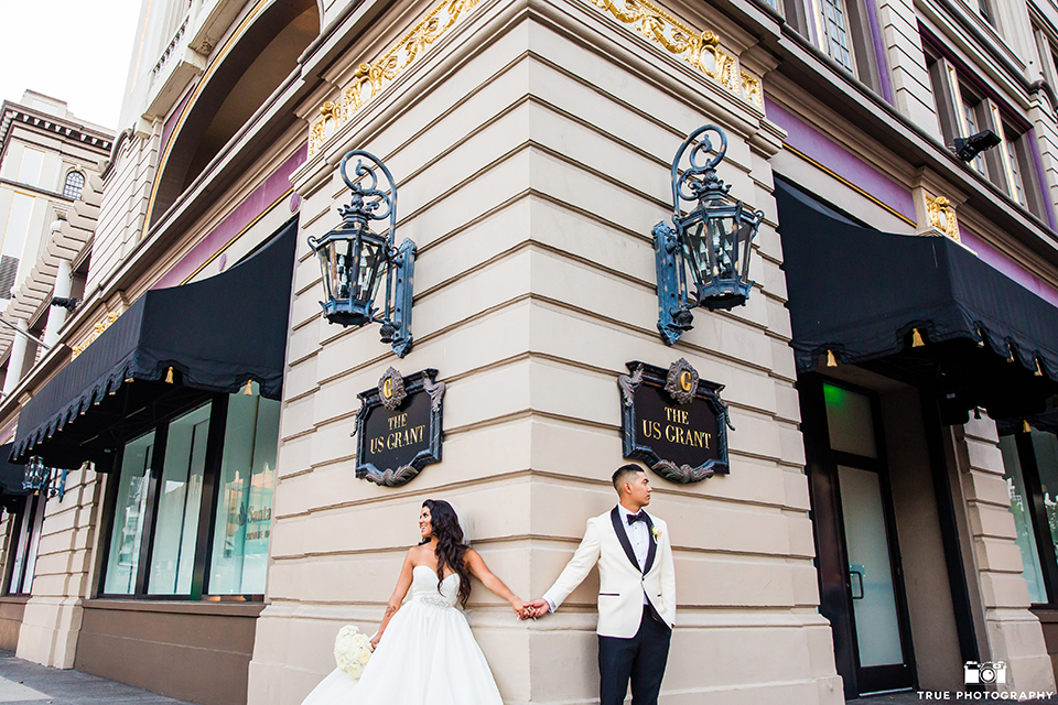 San diego glamorous wedding at the us grant hotel bride strapless ball gown with a tulle skirt and sweetheart neckline and a crystal belt with a long veil and groom white tuxedo jacket with a black shawl and black pants with a black bow tie and white floral boutonniere holding hands outside hotel