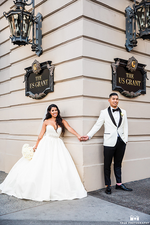 San diego glamorous wedding at the us grant hotel bride strapless ball gown with a tulle skirt and sweetheart neckline and a crystal belt with a long veil and groom white tuxedo jacket with a black shawl and black pants with a black bow tie and white floral boutonniere holding hands and smiling