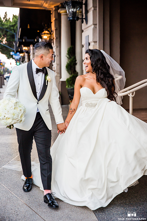 San diego glamorous wedding at the us grant hotel bride strapless ball gown with a tulle skirt and sweetheart neckline and a crystal belt with a long veil and groom white tuxedo jacket with a black shawl and black pants with a black bow tie and white floral boutonniere holding hands and walking