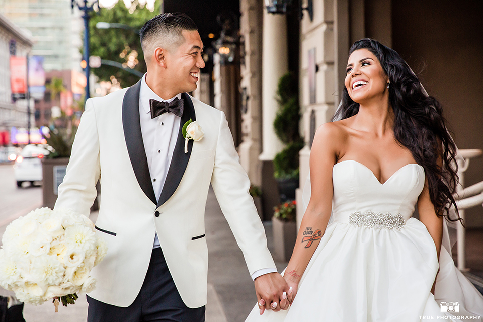 San diego glamorous wedding at the us grant hotel bride strapless ball gown with a tulle skirt and sweetheart neckline and a crystal belt with a long veil and groom white tuxedo jacket with a black shawl and black pants with a black bow tie and white floral boutonniere holding hands and walking smiling